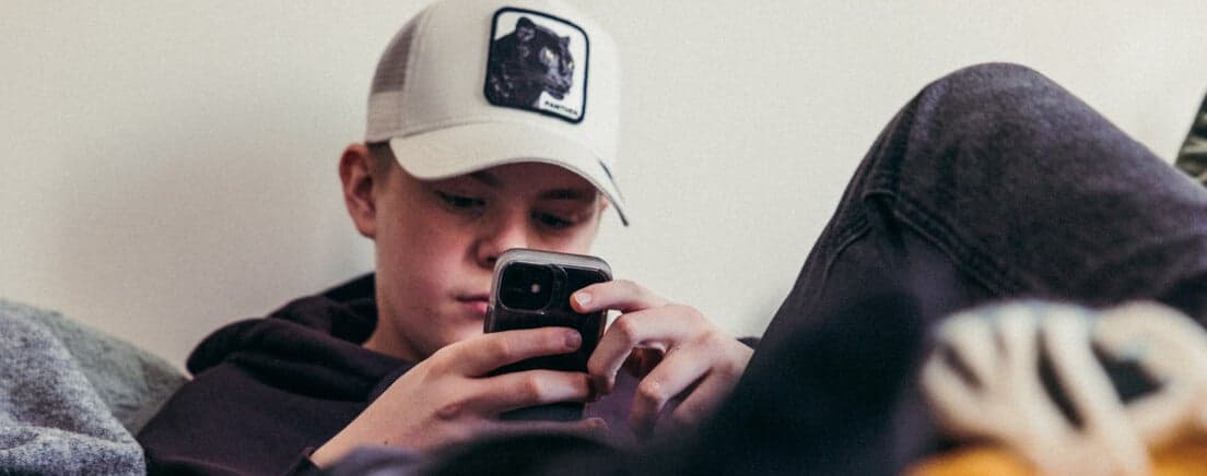 Pojke med mobil Pojke i keps ligger på en säng och tittar på sin mobil