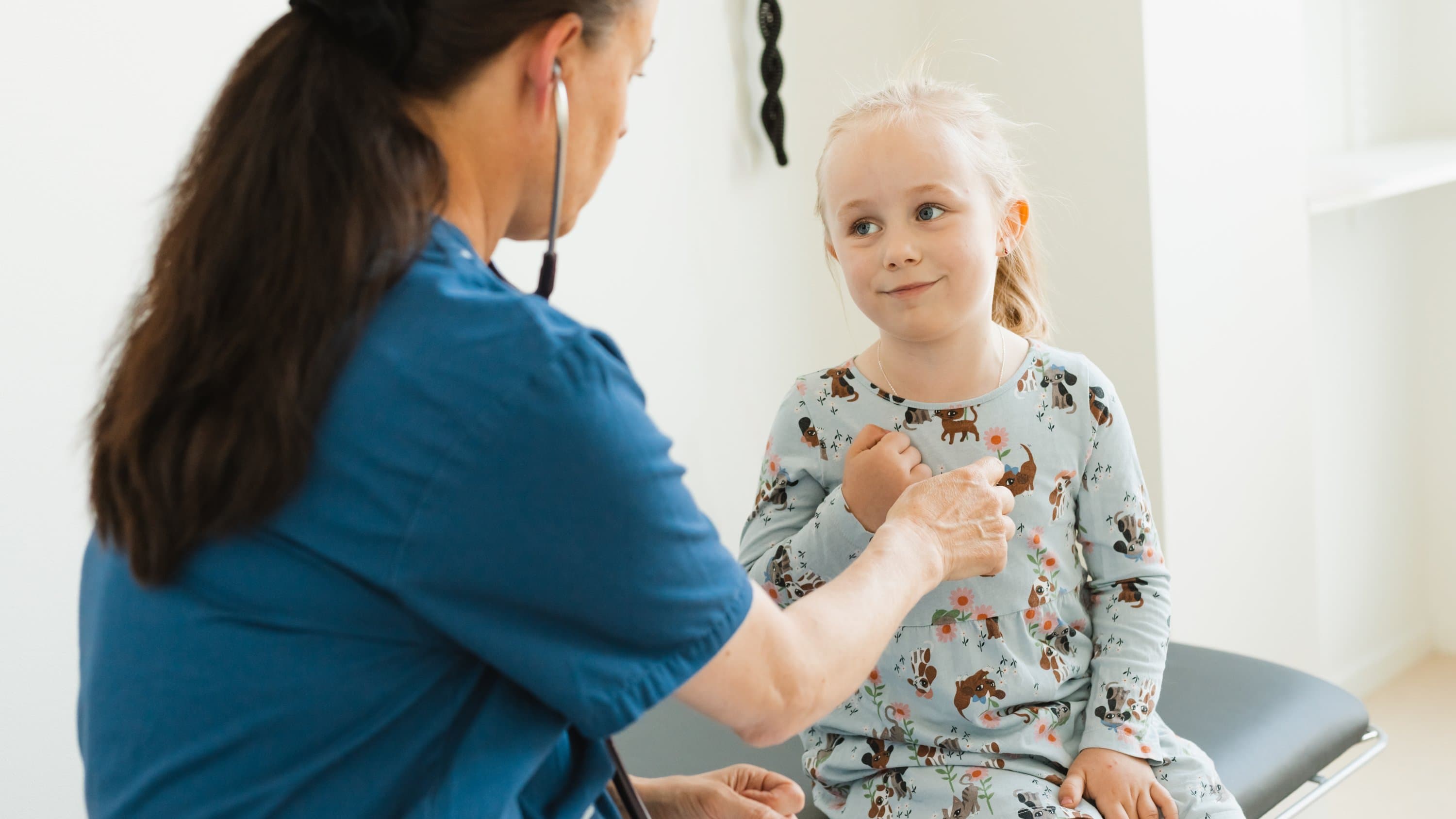 Läkare undersöker barn Kvinnlig läkare lyssnar på hjärtat på en liten blond flicka i mönstrad klänning