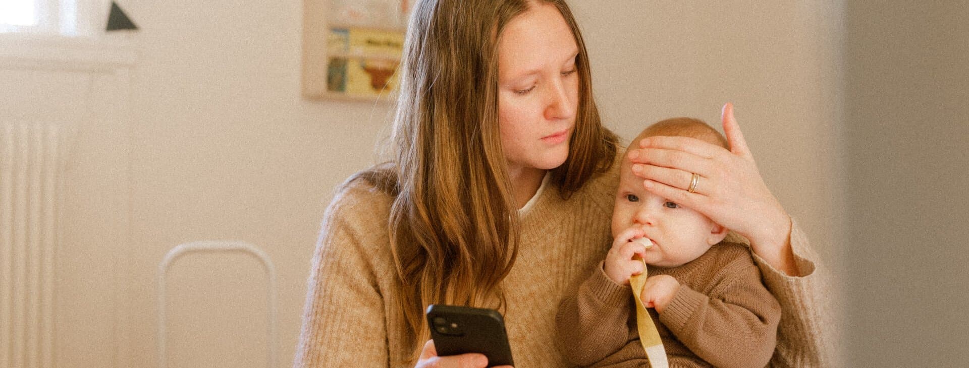 Mamma med bebis i knät håller telefon i ena handen och känner med andra handen på barnets panna.